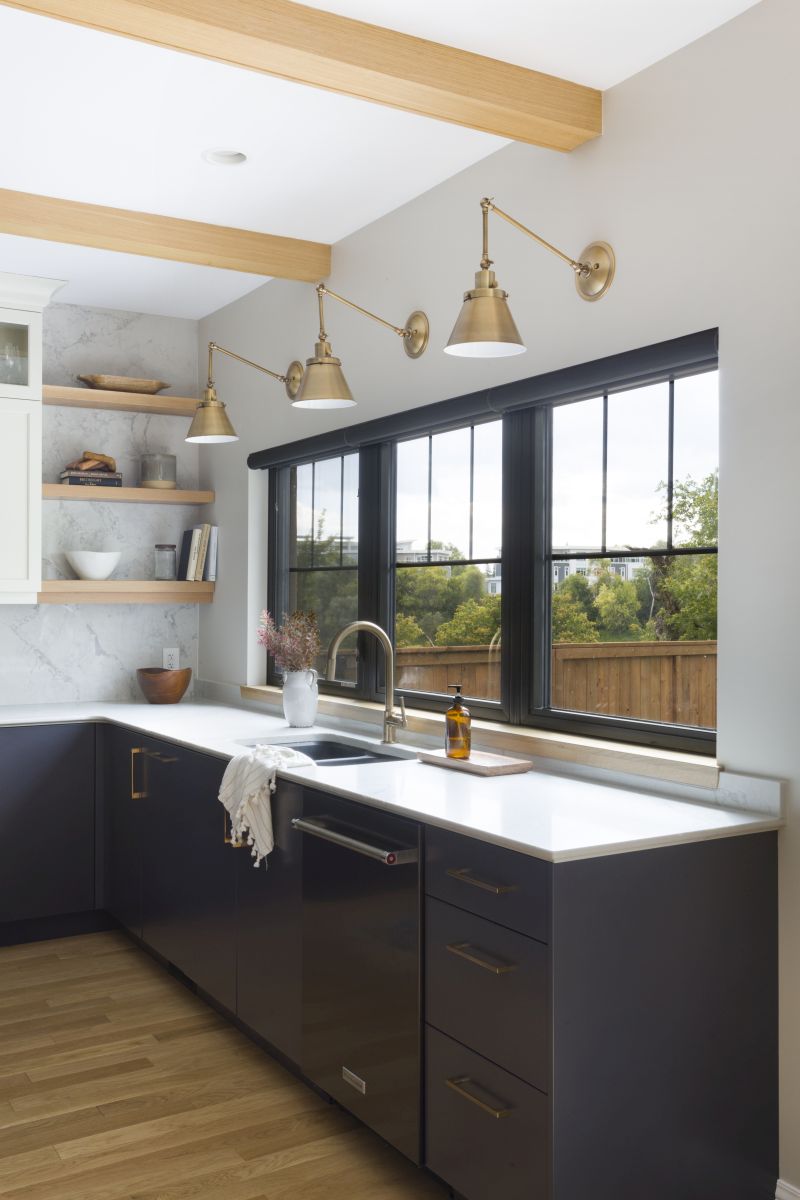 Modern kitchen with large black-trimmed windows, brass wall sconces, open wood shelving, and blue base cabinets with white countertops