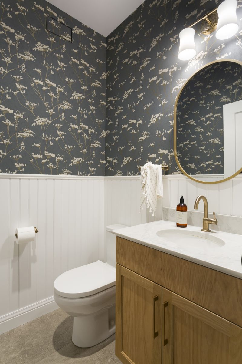 Transitional bathroom design featuring patterned wallpaper, beadboard wainscoting, natural wood vanity, and round mirror